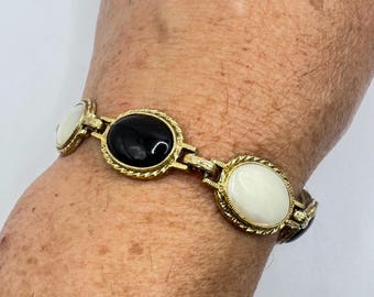 Pode incluir: Pulseira dourada com pedras ovais pretas e brancas. A pulseira tem uma borda texturizada em forma de corda em volta de cada pedra e é conectada por elos retangulares. A pulseira é usada no pulso.