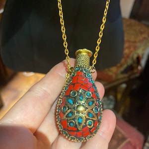 May include: A gold chain necklace with a red and turquoise pendant shaped like a bottle. The pendant is decorated with intricate details and a gold cap.