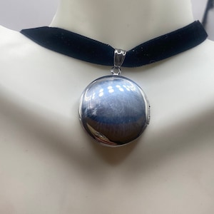May include: A silver locket pendant on a black velvet choker necklace. The locket is round and has a shiny, reflective surface.