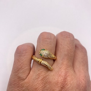 Puede incluir: Un anillo de serpiente de oro con piedras transparentes y verdes. La serpiente está enroscada alrededor del dedo y tiene un cuerpo texturizado.