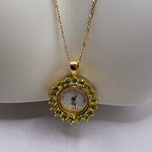 May include: A gold-toned necklace featuring a pendant watch. The watch face is white with black hands and hour markers, surrounded by a halo of green gemstones. The chain is a delicate, linked design.