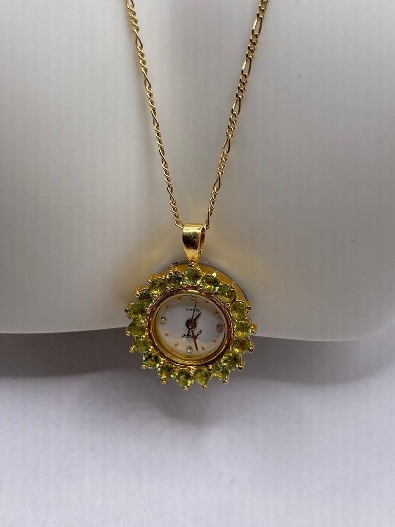 May include: A gold-toned necklace featuring a pendant watch. The watch face is white with black hands and hour markers, surrounded by a halo of green gemstones. The chain is a delicate, linked design.
