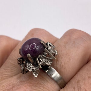 May include: A silver ring featuring a large, round, purple gemstone set in a textured, abstract design.