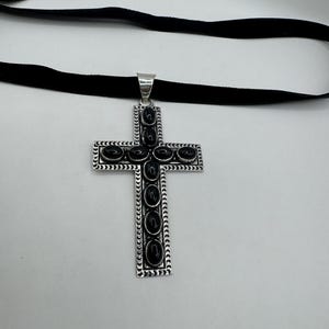 Op de afbeelding: Een zilveren kruis hanger met zwarte onyx stenen ingelegd in een decoratieve rand. De hanger is bevestigd aan een zwarte fluwelen choker.