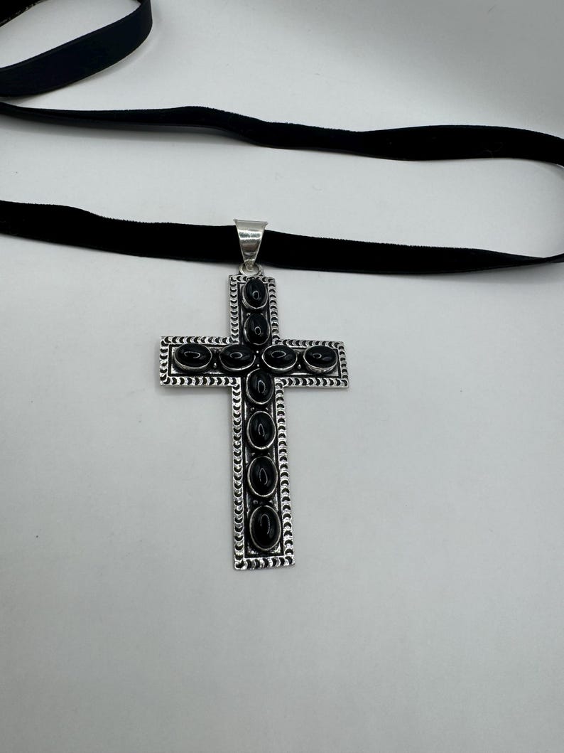 Op de afbeelding: Een zilveren kruis hanger met zwarte onyx stenen ingelegd in een decoratieve rand. De hanger is bevestigd aan een zwarte fluwelen choker.