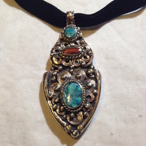 May include: A silver pendant necklace with a turquoise and red stone design. The pendant is ornate and features intricate details. The necklace is on a black velvet cord.