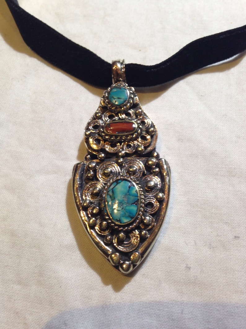 May include: A silver pendant necklace with a turquoise and red stone design. The pendant is ornate and features intricate details. The necklace is on a black velvet cord.