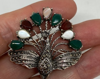 Puede incluir: Un broche de pavo real de plata con piedras verdes, rojas y blancas. El pavo real está posado en una rama y tiene un diseño detallado.