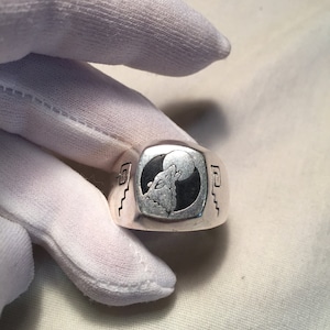 May include: A silver ring with a black and white design of a wolf howling at the moon. The ring has a square shape and a textured surface.