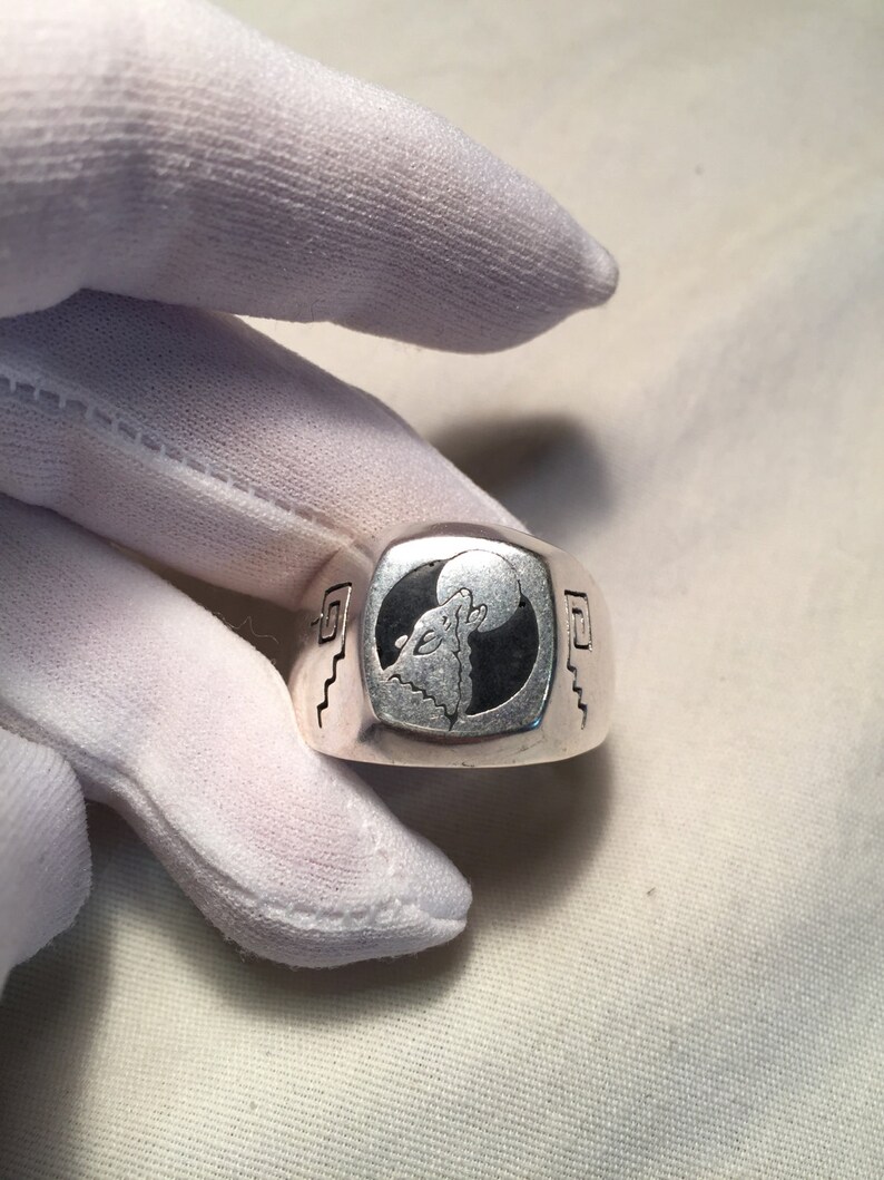 May include: A silver ring with a black and white design of a wolf howling at the moon. The ring has a square shape and a textured surface.