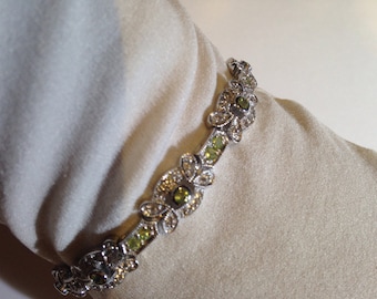 May include: Silver-toned bracelet with a floral design, featuring small, round, green gemstones and clear crystals. The bracelet is set against a soft, off-white background, highlighting its intricate details and elegant style.