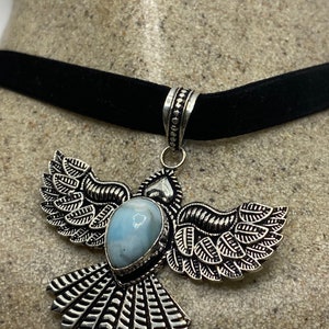 May include: A silver bird pendant with a blue gemstone set in the center, hanging from a black velvet choker.