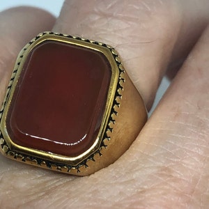 May include: A gold ring with a large, rectangular, red carnelian gemstone set in a gold bezel. The ring has a decorative, textured band.