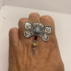 Puede incluir: Un anillo de mariposa de plata con piedras preciosas azules, rojas y amarillas. El anillo está decorado con intrincados diseños florales.