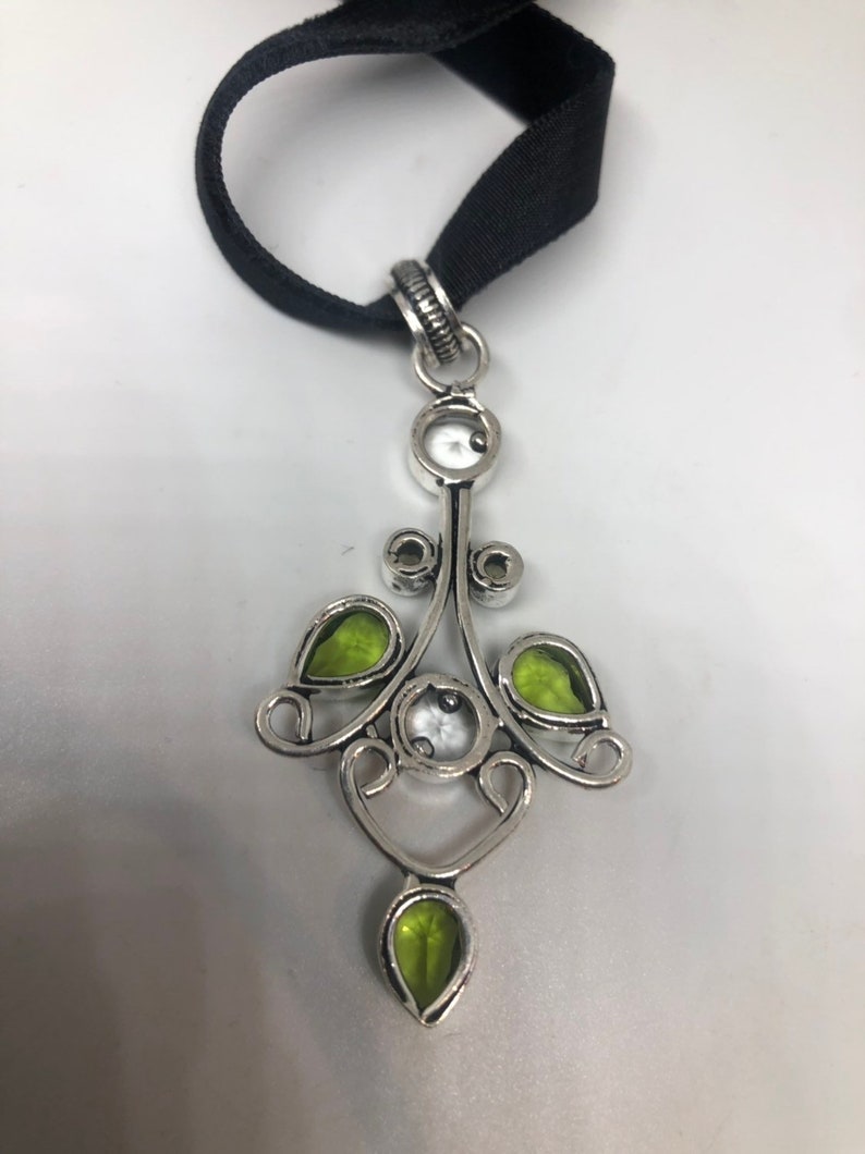 May include: A silver pendant necklace with a black ribbon. The pendant features a delicate design with green and white gemstones.