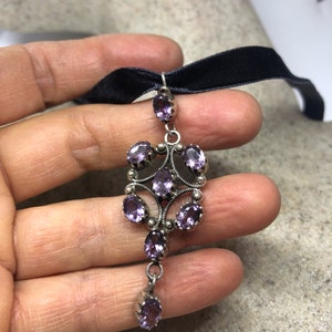May include: A silver pendant with a cluster of purple amethyst stones set in a filigree design. The pendant hangs from a black velvet ribbon.