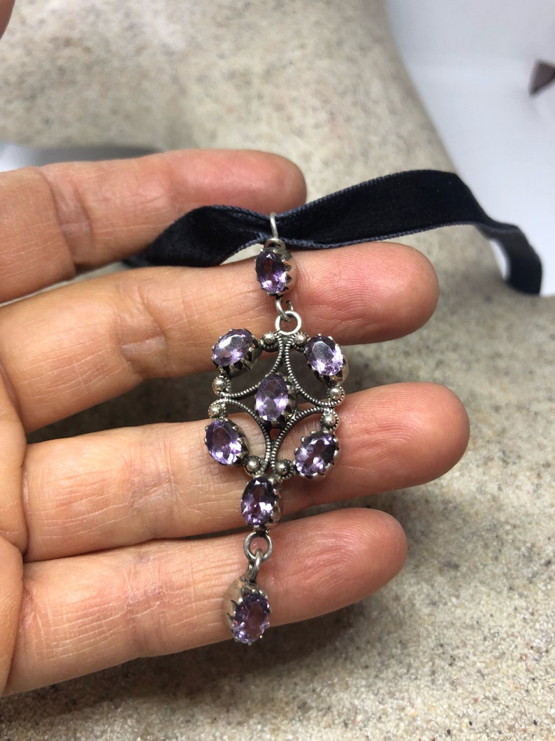 May include: A silver pendant with a cluster of purple amethyst stones set in a filigree design. The pendant hangs from a black velvet ribbon.