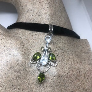 May include: A silver pendant necklace with a black velvet choker. The pendant features a white gemstone surrounded by green gemstones in a delicate, ornate design.