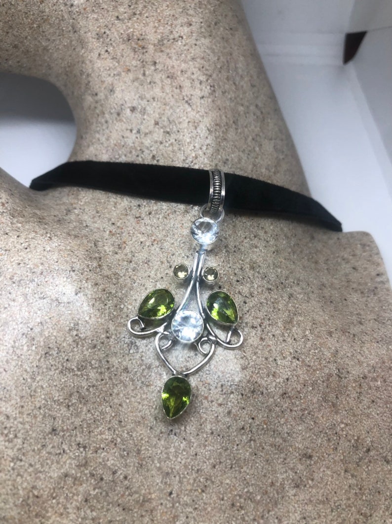 May include: A silver pendant necklace with a black velvet choker. The pendant features a white gemstone surrounded by green gemstones in a delicate, ornate design.