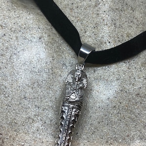 May include: A silver pendant necklace with a detailed Egyptian pharaoh design. The pendant is attached to a black velvet choker.