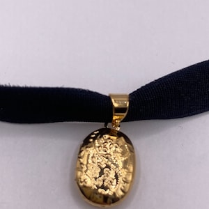 May include: A gold oval locket pendant with an intricate design hangs from a black velvet choker.
