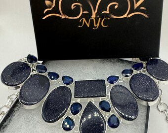 Puede incluir: Un collar de plata con una pieza llamativa que presenta múltiples piedras preciosas de color azul oscuro. El collar se muestra en una caja negra con el texto "Nemesis NYC" en la tapa.