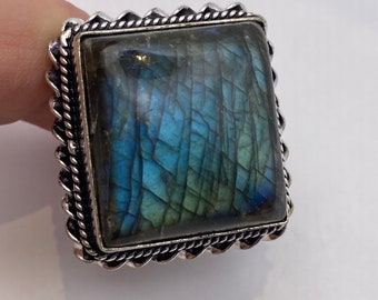 May include: A square silver ring with a large, iridescent blue and green labradorite gemstone set in a silver bezel. The ring has a decorative, rope-like border.