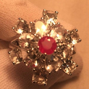 May include: A silver ring featuring a large red gemstone surrounded by smaller clear gemstones.