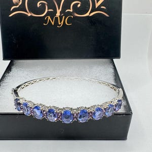 May include: A silver bangle bracelet with a row of oval-shaped blue gemstones set in a silver band. The bracelet is displayed in a black box with the text "Nemesis NYC" on the lid.
