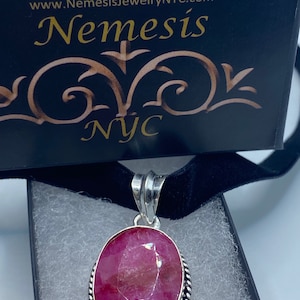 May include: A silver pendant with a large, oval-shaped, deep red gemstone. The gemstone is set in a silver bezel with a twisted rope design. The pendant hangs from a black velvet cord. The box has the text "Nemesis NYC".