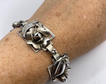 May include: A silver bracelet featuring a floral design with roses and leaves. The bracelet is made of a shiny, metallic material and is connected by hinges. The floral elements are intricately detailed, showcasing the craftsmanship of the piece.
