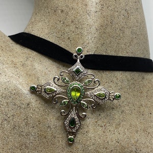 Puede incluir: Un colgante de cruz de plata con piedras preciosas verdes y blancas. El colgante está en una gargantilla de terciopelo negro.