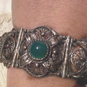Puede incluir: Un brazalete de plata con intrincados detalles de filigrana y acentos de piedras preciosas verdes.