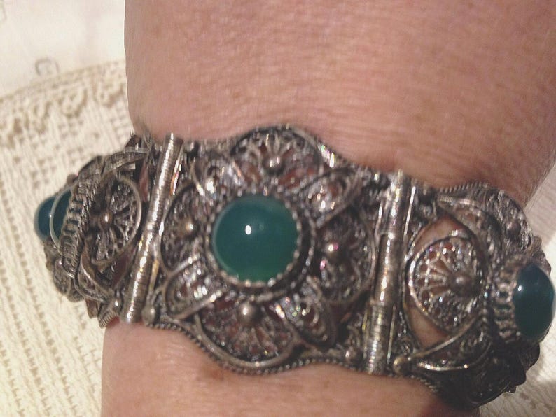 Puede incluir: Un brazalete de plata con intrincados detalles de filigrana y acentos de piedras preciosas verdes.