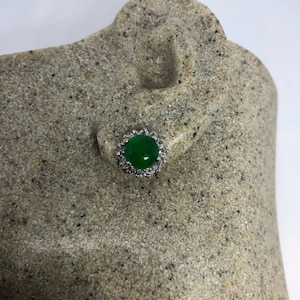 Op de afbeelding: Een groene edelsteen oorbel met een zilveren zetting en een decoratieve rand van kleine heldere stenen.