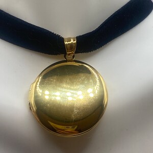 May include: A gold locket pendant on a black velvet choker necklace. The locket is round and has a shiny, polished finish.