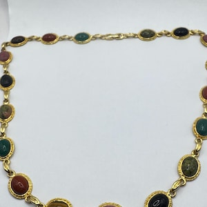 May include: Gold-toned necklace featuring oval-shaped gemstones in various colors, including red, green, black, and pink. The gemstones are set in a gold-colored metal frame and linked together with a delicate chain. A vintage-inspired jewelry piece.
