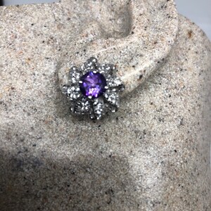 May include: A silver earring with a purple gemstone surrounded by smaller clear gemstones in a flower shape.