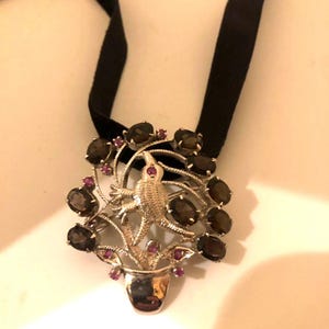 May include: A silver pendant necklace with a bird design and brown and pink gemstones. The necklace is on a black ribbon.