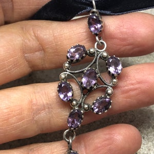 May include: A silver pendant with a delicate design featuring multiple amethyst gemstones. The pendant is attached to a black velvet ribbon.