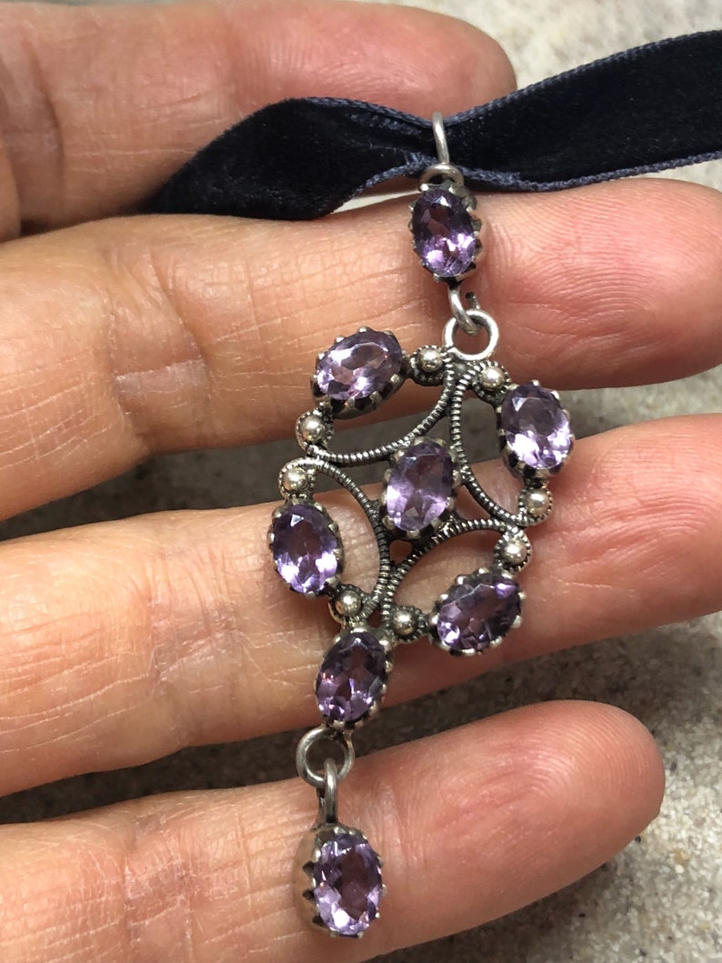 May include: A silver pendant with a delicate design featuring multiple amethyst gemstones. The pendant is attached to a black velvet ribbon.