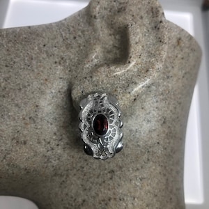 May include: A silver-toned earring with an oval, dark red gemstone at its center. The earring features an ornate, filigree design with small, clear accent stones. The earring is displayed on a gray, textured surface.