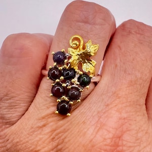 May include: A silver ring featuring a cluster of dark red and green gemstones, resembling grapes. The ring has a gold-colored leaf and stem design. The ring is worn on a finger.
