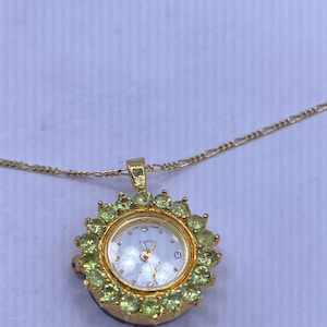 May include: A gold-toned pendant watch necklace. The watch face is white with gold hands and small diamond accents. The watch is surrounded by green gemstones and hangs from a delicate gold chain.