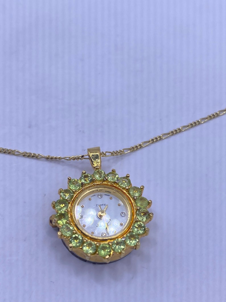 May include: A gold-toned pendant watch necklace. The watch face is white with gold hands and small diamond accents. The watch is surrounded by green gemstones and hangs from a delicate gold chain.