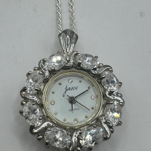 Op de afbeelding: Zilverkleurig hangerhorloge met een witte wijzerplaat en de merknaam "Jasco". Het horloge is omringd door heldere, ovale edelstenen en hangt aan een zilveren ketting. De wijzerplaat toont het woord "Quartz" en "Japan".