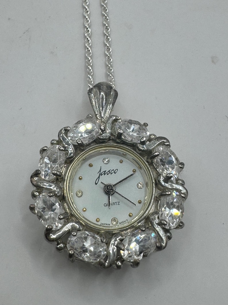 Op de afbeelding: Zilverkleurig hangerhorloge met een witte wijzerplaat en de merknaam "Jasco". Het horloge is omringd door heldere, ovale edelstenen en hangt aan een zilveren ketting. De wijzerplaat toont het woord "Quartz" en "Japan".