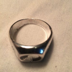 May include: A silver signet ring with a smooth, rounded surface. The ring has a raised design on the top.