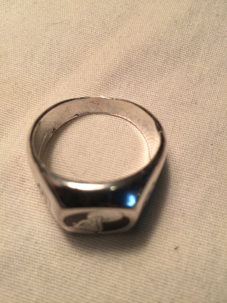 May include: A silver signet ring with a smooth, rounded surface. The ring has a raised design on the top.
