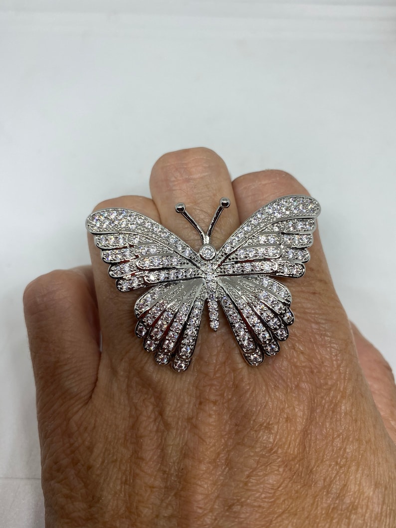 K&ouml;nnte beinhalten: Ein silberner Schmetterlingsring mit klaren Zirkonia-Steinen. Der Ring ist gro&szlig; und hat ein detailliertes Design.